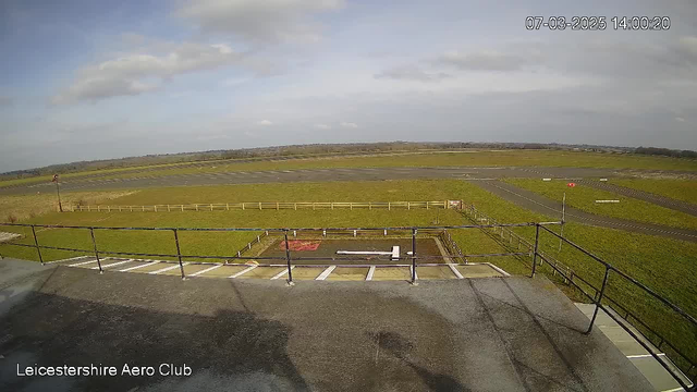 A view from a high vantage point at Leicestershire Aero Club, showing a large grassy area and a runway. The sky is partly cloudy with patches of blue. In the foreground, there is a railing along the edge of the platform. The runway is visible in the background, with a few planes parked and a red windsock on the left side. There is a wooden fence bordering the grassy area, and markings on the runway indicate the taxiway and runway layout. The time displayed is 14:00 on March 7, 2025.