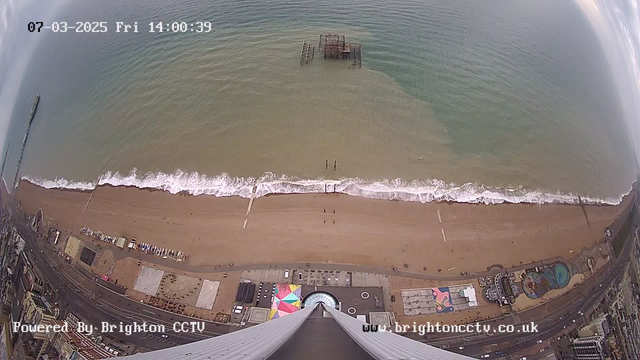 A panoramic view from above a beach with a sandy shoreline meeting the ocean. Gentle waves are lapping at the shore, with some beachgoers visible in the distance. An old pier stretches into the water, partially submerged. There are colorful beach structures and a circular pattern on the ground below, along with several parked vehicles near the road. The sky is partly cloudy, and the image timestamp indicates it is Friday, March 7, 2025, at 2:00 PM.