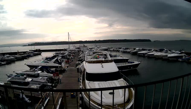 A view of a marina filled with moored boats and yachts, with a cloudy sky above. The image captures a wooden dock lined with various vessels, some of which are larger motorboats. The water is calm, and there are distant land formations visible on the horizon. The overall atmosphere appears tranquil yet overcast.