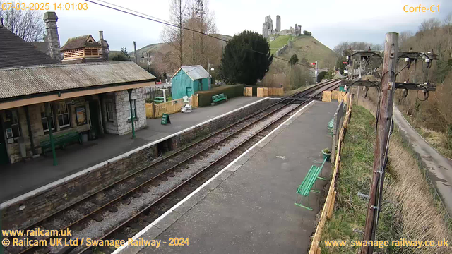 A view of a railway station with stone buildings and a wooden platform. There are green benches and a wooden fence surrounding the area. In the background, a hill features a castle ruin. The tracks are visible, running parallel to the platform, and a power pole with wires stands on the right side of the image. The weather appears overcast.