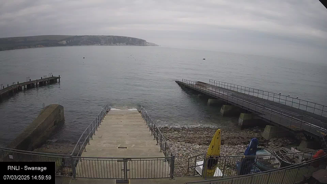 A cloudy day at a seaside location shows calm waters with a few small boats visible. In the foreground, there is a concrete ramp leading down to the water, bordered by a metal railing. To the right, two piers extend into the sea, one with a gently sloping surface. At the base of the ramp, there are rocks and pebbles scattered along the shoreline. A yellow kayak is propped up against the railing, along with other boats partially visible. Soft waves lap against the shore.