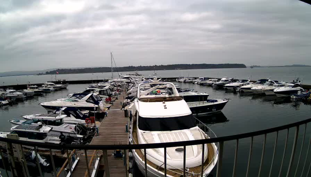 A marina filled with numerous boats moored along a wooden dock. The scene is dominated by a cloudy sky, with a calm body of water reflecting the boats and clouds above. Several motorboats and yachts are visible, varying in size and design. In the background, there are trees and a hilly landscape on the shore. The atmosphere appears tranquil, typical of a marina setting.