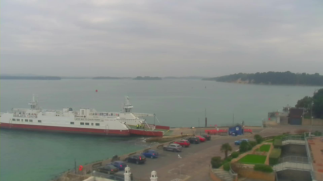 A ferry with a white and red hull is docked near a shoreline. The water is calm and reflects the overcast sky. Several parked cars, mainly in blue and red, are visible along the waterfront, and there are a few people near the ferry. In the background, a hilly landscape can be seen across the water, and there is a small structure on the shore. The scene conveys a quiet, cloudy day by the water.