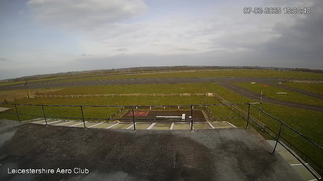 A view from a webcam positioned on a building overlooking an airfield. The foreground shows a flat rooftop area with a railing. Below, there is an expansive green field with patches of grass and some dirt. In the distance, there are two runways marked by dark asphalt, with a few scattered white lines. A sign is visible on a post near the edge of the runway. The sky is mostly cloudy, and the overall scene appears calm and open. The bottom left corner includes text identifying the location as "Leicestershire Aero Club," along with a timestamp.