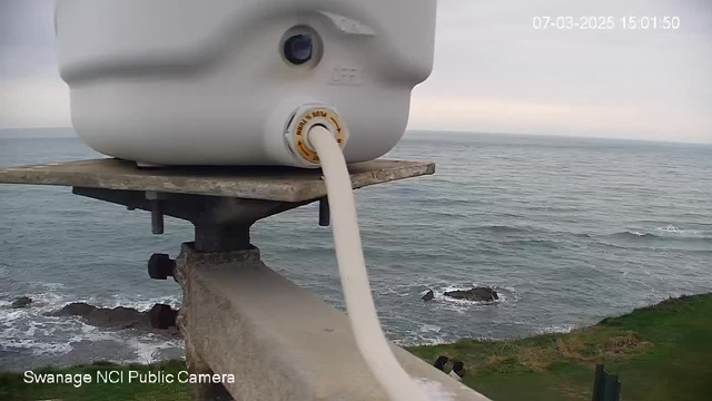 A close-up view of a white cylindrical object placed on a gray, metal surface, situated along the edge of a coastal area. The object has a small hole on its side and a yellow-labeled connector from which a hose extends down towards the ground. In the background, the ocean is visible with gentle waves, while the sky is overcast. There are rocky formations and a green grassy area near the shoreline. There are also a couple of small figures seen at the edge of the grass.