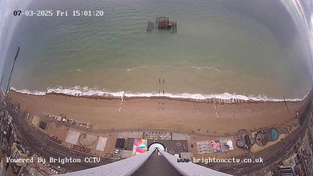 A bird's-eye view of a beach scene showing a sandy shoreline meeting the ocean. Light waves gently lap against the shore, with a deserted pier visible in the water. There are small figures along the beach, and the beach is lined with various amenities, including colorful beach huts and attractions. The sky shows some clouds, and the image includes a timestamp and logo at the bottom.