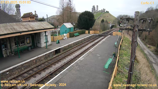 A view of a train station platform with green benches along the waiting area. On one side, there is a stone building with a sloping roof and a wooden section above it. A green shed is visible in the background, alongside a wooden fence surrounding a grassy area. In the distance, the ruins of a castle are on a hill. The train tracks extend into the image, leading toward the horizon. A power pole with wires stands on the right side of the frame. The sky is overcast.