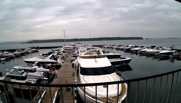 A view of a marina on a cloudy day, featuring numerous boats anchored in calm water. In the foreground, a large white yacht is docked alongside a wooden pier. Other boats of various sizes and colors are scattered throughout the marina. The background shows a hazy shoreline, and the sky is overcast with gray clouds.