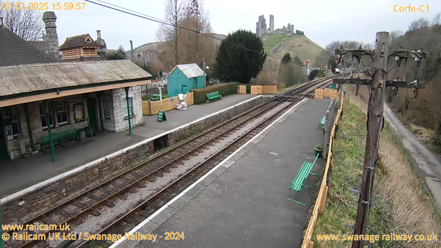 A view of a train station platform with a stone building on the left featuring a sloped roof and a wooden decorative element. Several green benches are visible on the platform, along with signs indicating "WAY OUT." In the background, a hill is partially obscured by trees, with ruins of a castle situated at the top. The tracks run alongside the platform, and there are telephone poles with electrical wires on the right side of the image. The sky appears cloudy.