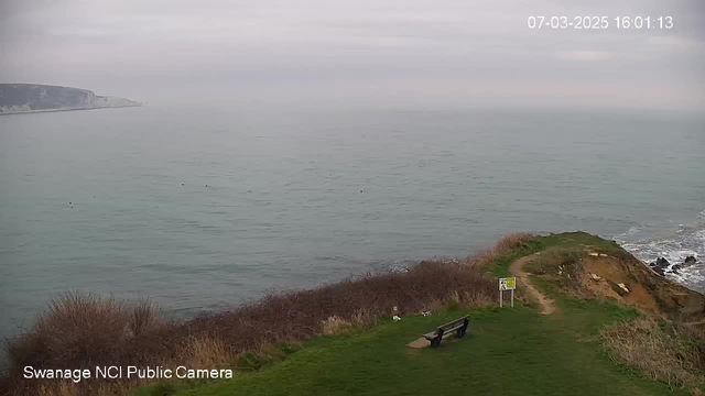 A coastal scene viewed from a grassy hillside. The foreground features a wooden bench and a path leading down toward the water's edge. In the background, a calm sea stretches out to the horizon under a cloudy sky. The cliffs can be seen in the distance to the left, and there are patches of shrubbery and grass on the hillside. The image indicates a cool, overcast day.