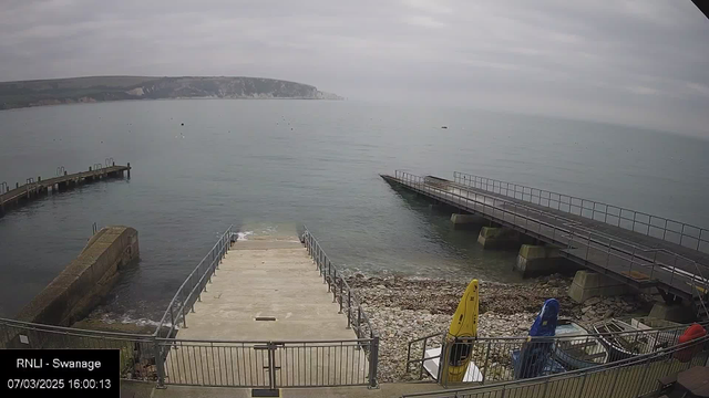 A coastal view showing a gray sky over calm waters. In the foreground, there are concrete steps leading down to the water, flanked by metal railings. To the left, a small pier extends into the sea, and there are scattered rocks along the shoreline. On the right, another pier is visible, sloping down into the water. Two colorful kayaks, one yellow and another blue, are stored near the edge, while a few small boats are scattered nearby. The background features a green hillside and white cliffs in the distance. The scene appears quiet and tranquil.