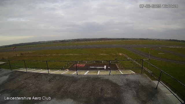 A view from a webcam at Leicestershire Aero Club shows a large expanse of green grass and a runway under a cloudy sky. In the foreground, there is a railing, with a walkway leading towards a small area that appears to have a platform and some markings. To the left, a windsock indicates wind direction. The background features distant trees and a horizon with more flat land.