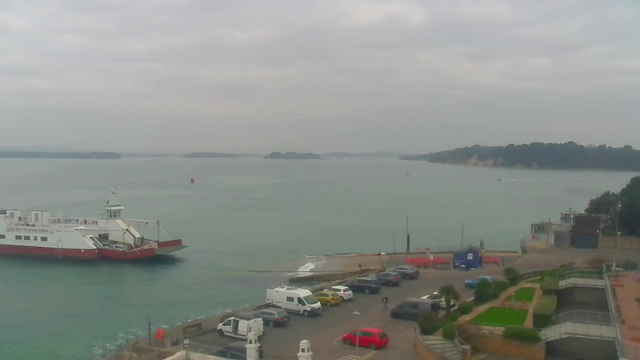 A coastal view featuring a ferry approaching a dock. The water is calm with a light turquoise hue, and several small boats can be seen in the distance. A concrete pier extends into the water, and there is a parking area filled with various vehicles, including cars and vans. The sky is overcast with gray clouds, and small islands are visible in the background. There is a grassy area with steps leading down towards the waterfront, along with some shrubs and palm-like plants.