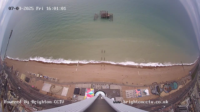 Aerial view of a beach with gently rolling waves and a sandy shore. There are a few small people walking along the water's edge. An old pier structure stands in the water, partially submerged. Along the beach, there are colorful amusement park structures and rides visible. The image captures a wide expanse of the sea and coastline, with some clouds in the sky. The date and time are displayed at the top.