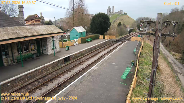 A view of a railway station with a stone building and a sloped grassy hill in the background featuring ruins. The platform is lined with several green benches and has a stone wall along the edge. Railway tracks run parallel to the platform, and a wooden fence encloses a small waiting area with a sign that says "WAY OUT." There are trees and bushes near the station, with a path visible on the right side of the image. The sky is overcast.