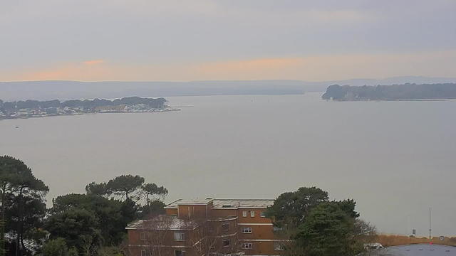 A view of a calm body of water under a cloudy sky. The horizon shows distant hills, while the foreground features a brown multi-story building surrounded by trees. To the left, a marina with several boats is visible along the shore. The overall scene is serene and slightly overcast, with soft light illuminating the landscape.