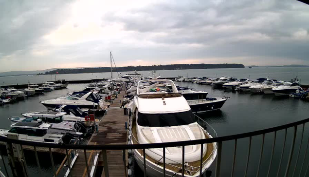 A cloudy scene at a marina featuring numerous boats docked in a calm body of water. In the foreground, a large white yacht is visible, alongside several other boats of various sizes and colors, including blues and grays. A wooden walkway extends along the dock, leading to the marina. In the background, green hills are partially shrouded in clouds. The sky appears overcast, suggesting potential rain.