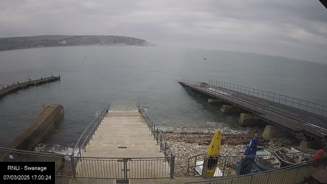 A view of a calm seaside with a cloudy sky, featuring a concrete ramp leading down to the water. On the left, a rocky pier extends into the sea, and on the right, there is a larger wooden pier with railings. Several small boats are visible on the water, and some yellow and blue kayaks are positioned near the shore, resting on a gravel area.