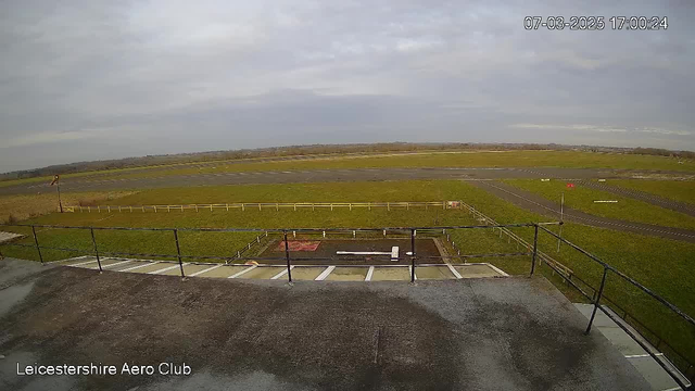 A view from a webcam at the Leicestershire Aero Club. The image shows a grassy area with a long, straight runway in the foreground. There is a wooden fence surrounding part of the lawn area. In the distance, the sky is mostly cloudy, with hints of blue peeking through. The time displayed is 17:00 on March 7, 2025.