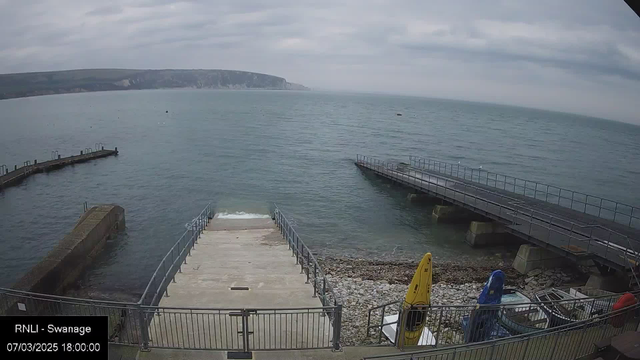 A calm coastal scene featuring the sea under a cloudy sky. In the foreground, there is a concrete staircase leading down to the water, flanked by railings. To the right, there are two piers extending into the water, one with a ramp and the other with a railing. On the shore, a sandy and pebbly area is visible, alongside yellow and blue kayaks. The distant hills and cliffs create a scenic backdrop.