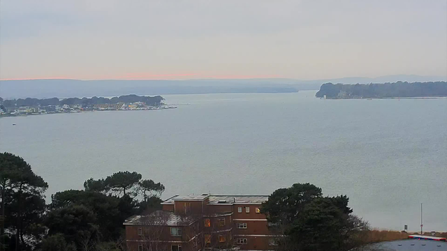 A panorama of a calm body of water stretches across the scene, with a faint horizon in the distance. On the left, there is a waterfront with small buildings and some greenery, transitioning to boats docked at a marina. In the foreground, trees are visible, adding contrast to the view, while the sky appears overcast with soft gray tones, hinting at a low light environment. The water reflects these subdued colors, creating a serene atmosphere.