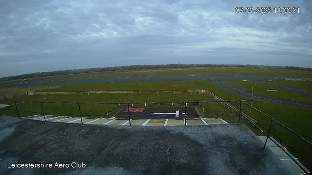 A view from a webcam at Leicester Aero Club showing a grassy airfield under a mostly cloudy sky. The scene includes a runway area with a small section of tarmac in the foreground, surrounded by a wooden fence. There are light poles and ground markings visible on the runway. The date and time are displayed in the top right corner, indicating March 7, 2025, at 18:00:21.