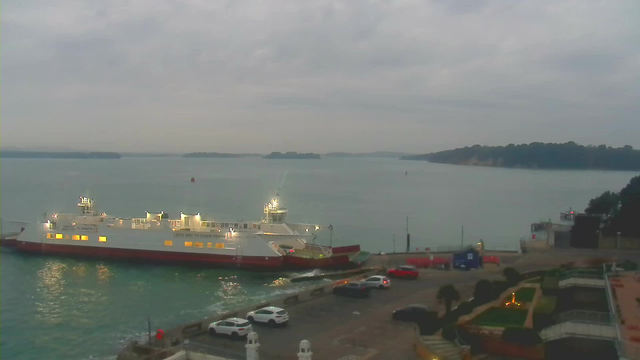 A view of a calm body of water under a cloudy sky, with a large ferry boat docked at the shore. The ferry is illuminated and has a red and white design. In the foreground, several parked cars are visible along the waterfront, and there is a landscaped area with plants and pathways. In the background, there are distant islands and a hint of land along the shoreline.