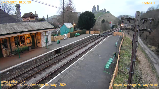 A railway station scene with a stone building featuring a sloping roof and several large windows, situated at the left side of the image. There are green benches on the platform and a sign reading "WAY OUT." The platform is bordered by a wooden fence. In the background, a hill is visible with the remains of a castle at the top, surrounded by sparse trees and shrubbery. The tracks run parallel to the platform, extending into the distance. The setting appears to be during twilight, with a faint light in the sky.