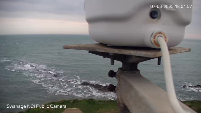 A webcam is mounted on a metal stand, capturing a view of the sea. The foreground features a white cylindrical device with a visible power cord connected to it. In the background, waves gently flow over rocky formations in the water, with a cloudy sky overhead. The scene is slightly gloomy, suggesting it may be early evening.