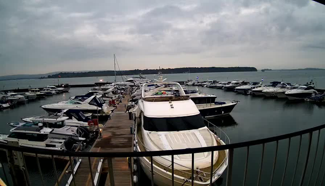 A marina scene featuring multiple boats docked along a wooden pier. In the foreground, a large white yacht is moored, with several smaller boats around it, some of which are blue and white. The water is calm and reflective, with a cloudy sky overhead and hints of land visible in the distance. The image captures the tranquil atmosphere of the marina.