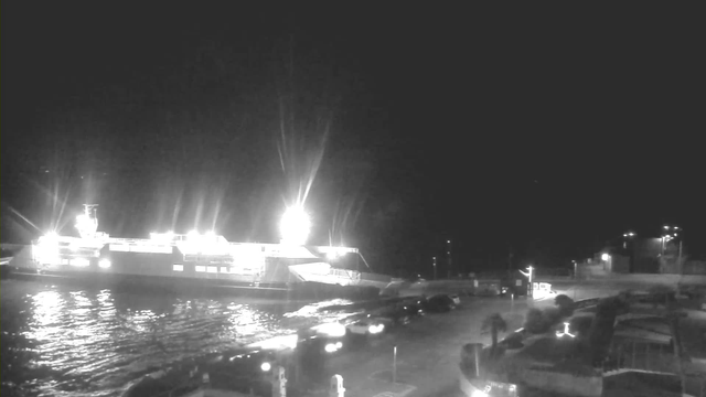 A brightly lit ferry docked at a dark marina. The ferry is illuminated from multiple angles, creating reflections on the water. In the foreground, several vehicles are parked along the edge of the marina. The background features distant lights and structures, suggesting additional buildings or a loading area. The scene has a nighttime ambiance with a mostly dark sky.