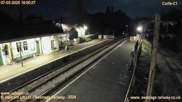 A nighttime view of a railway station. The platform is empty, illuminated by overhead lights. To the left, there is a stone building with large windows, featuring a green bench outside. A sign indicates "WAY OUT." On the right side of the image, there are several green benches along the platform, with a wooden fence and a lamp post visible in the background. The railway tracks run through the center, leading into the darkness. The sky is dark, and a few faint outlines of buildings are visible in the background.