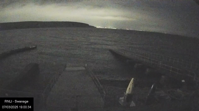 A dark, nighttime scene of a body of water with rippling waves. In the foreground, there is a wooden dock that extends into the water. To the right, two small boats with pointed bows are positioned near the shore. In the distance, a faint outline of land can be seen across the water, with a cloudy sky overhead and some distant lights visible on the horizon. The overall mood is tranquil with a sense of calmness.