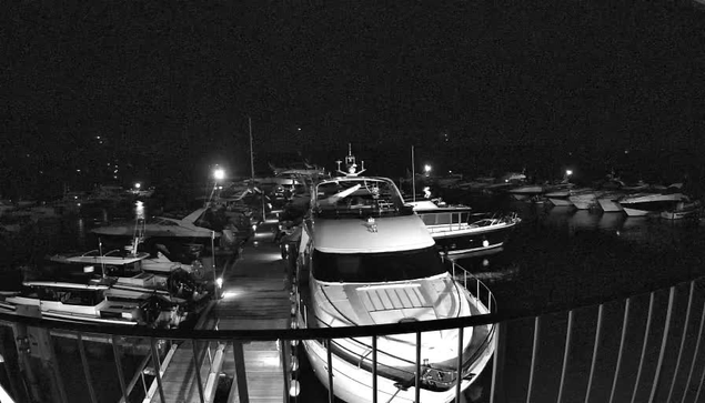 A nighttime view of a marina filled with boats. The scene is dimly lit, with several boats docked along wooden walkways. A large yacht is prominently positioned in the foreground, and various smaller boats are visible in the background. Some lights illuminate the area, reflecting off the water, creating a serene but shadowy atmosphere.