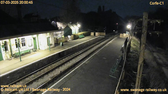 A nighttime view of a railway station platform. The platform is illuminated by overhead lights, showing a wooden bench on the left side and a green bench on the other side. In the background, there are buildings with a warm glow from windows. The railway tracks are visible, leading off into darkness. There is a wooden fence in the distance with a "WAY OUT" sign facing the viewer. The atmosphere is quiet and tranquil.