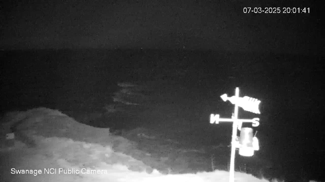 A dark, nighttime scene showing a large body of water, possibly the sea. In the foreground, there is a weather vane on a pole, positioned at the center right. The weather vane includes a fish design and is oriented towards the north. The coastline is faintly visible along the bottom left, while the water appears calm with slight ripples. The image is predominantly dark with minimal visible detail. In the upper right corner, there is a timestamp indicating the date and time.