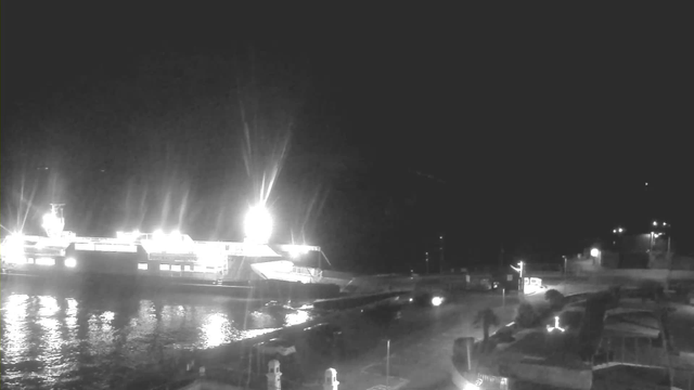 A nighttime view of a harbor with a large illuminated ferry docked on the left side of the image. The water reflects the bright lights from the ferry and other light sources on the shore. Various boats are visible in the foreground, with some lights and structures along the waterfront, creating a sense of activity in the area. The scene is predominantly dark with contrasting bright elements.