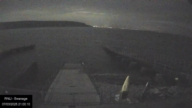 A dimly lit coastal scene at night, featuring a calm body of water reflecting faint light from the clouds above. In the foreground, there are a few structures, including a wooden pier extending into the water. Two kayaks, one yellow and one blue, are positioned beside the pier, hinting at a recreational area. The background showcases a distant shoreline with scattered lights, indicating a town or city. The atmosphere is serene, with a few clouds obscuring the moonlight, creating a tranquil nighttime setting.