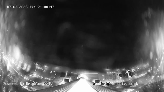 A black and white image captured from a high viewpoint at night. The foreground features a tall building with a pointed top, possibly a spire. Below, the ground is illuminated by various streetlights, creating a pattern of light along the edges. Buildings and structures are visible in the lower part of the image, with some blurred areas suggesting moisture or reflections on the camera lens. The date and time are displayed in the upper left corner, indicating it is Friday, March 7, 2025, at 9:00 PM.