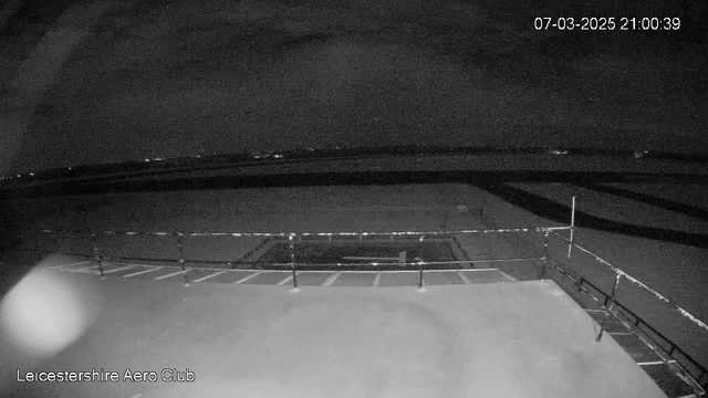 A dimly lit aerial view of an airport runway and surrounding area at night. The foreground shows a flat rooftop with a railing. There are faint lights visible in the distance, suggesting nearby buildings or vehicles. The overall scene is dark, indicating it is nighttime. In the top right corner, the date and time are displayed: 07-03-2025, 21:00:39. The image has a grainy quality typical of low-light conditions.