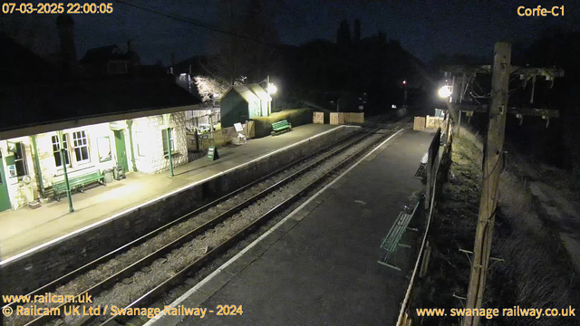 A dimly lit train platform at night is depicted. The platform features stone walls and green benches, with a small building having large windows and a sign that reads "WAY OUT." Two railway tracks run parallel, with gravel in between. On the right, a wooden pole with power lines is visible, along with a single light illuminating the area. There are faint outlines of trees in the background, contributing to the nighttime atmosphere. The overall scene conveys a quiet and peaceful waiting area at a rural train station.