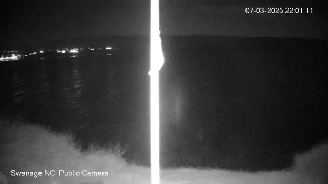 A dimly lit night scene showing a calm sea with small lights visible on the distant shoreline. In the foreground, there is a bright vertical pole, and the date and time in the corner indicate "07-03-2025 22:01:11." The overall atmosphere is dark with soft reflections on the water.