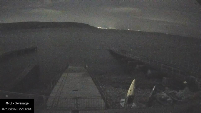 A dimly lit scene depicting a dock area at night with water visible in the foreground. The dock is made of wood and extends into the water, while a few boats, including a kayak, are seen on the ground near the dock. In the distance, faint lights are visible along the shoreline, suggesting an area far from the dock. The scene conveys a calm and serene atmosphere with clouds in the sky.