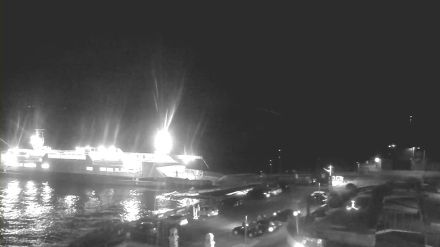 A nighttime scene featuring a brightly lit ferry docked at a harbor. The water reflects the lights of the ferry and surrounding structures. There are several vehicles parked near the harbor, and faint lights from the shore illuminate the area. The background is mostly dark, emphasizing the illuminated ferry and nearby elements.