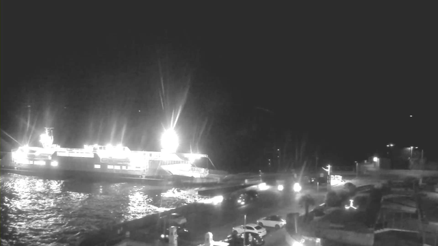 The image shows a dark nighttime scene featuring a large illuminated ferry docked at a harbor. The ferry is lit up brightly, with reflections glimmering on the water's surface. There are several parked cars in the foreground, and faint lights can be seen along the shore, indicating buildings and streetlights. The overall ambiance is quiet and calm, typical of a nighttime waterfront setting.