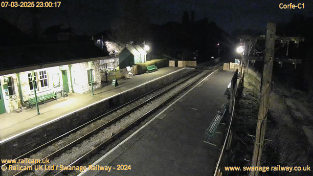 A dark scene at a railway station during the night. The platform is mostly deserted with a few benches visible. To the left, there is a small building with a light illuminating the entrance, which features several windows. A sign indicating "Way Out" can be seen. The railway tracks run along the bottom of the image, and there is minimal lighting from the lamps along the platform. The surrounding area appears quiet, with trees and bushes visible in the background.