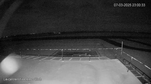 A dimly lit view from a webcam showing the edge of a rooftop with a railing. In the background, a dark landscape is visible, possibly indicating fields or roads, with faint lights scattered in the distance. The time stamp in the corner indicates the image was taken on March 7, 2025, at 23:00:33. The quality is low with a grainy appearance, typical of nighttime visibility.