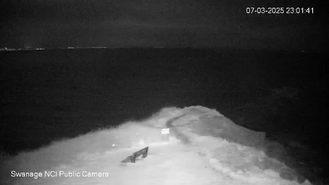 A nighttime view of a rocky shoreline with an expanse of dark water in the background. A small sign is positioned near the edge of the water, and a section of the shore appears to have a path leading toward it. The scene is illuminated with minimal light, creating a dark and moody atmosphere.