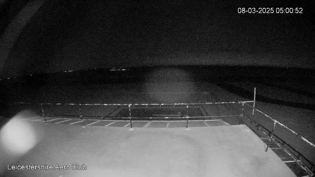 A dimly lit scene from a webcam showing a flat surface with a railing in the foreground. The background is mostly dark, with faint distant lights suggesting the presence of a body of water or landscape. The date and time are displayed in the top right corner.