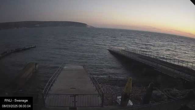 A dock with a concrete walkway extends into a choppy sea under a lightening sky at dawn. On the left side, there are wooden pilings jutting into the water. In the background, a coastal cliff is visible, silhouetted against the soft hues of the morning light. The scene conveys a calm yet dynamic atmosphere as waves gently lap at the shoreline.